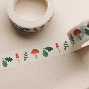 May include: A roll of white washi tape with a repeating pattern of red and white mushrooms, green leaves, and brown stems. The tape is unrolled and shows the pattern.