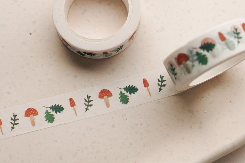 May include: A roll of white washi tape with a repeating pattern of red and white mushrooms, green leaves, and brown stems. The tape is unrolled and shows the pattern.