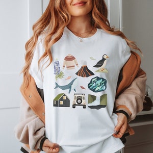 May include: White t-shirt with a colorful graphic design featuring an Icelandic landscape, including a puffin, a whale, a volcano, a house, a sweater, and a car.