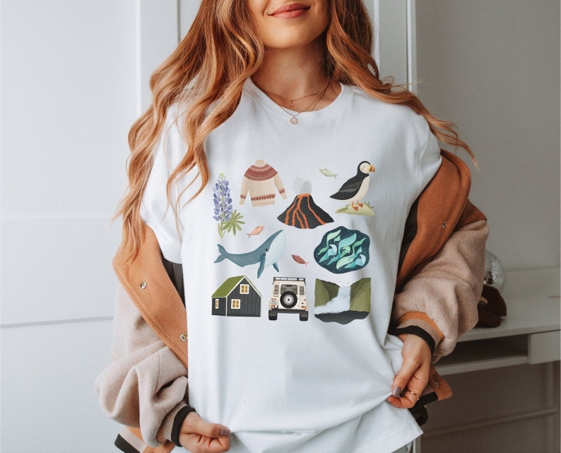 May include: White t-shirt with a colorful graphic design featuring an Icelandic landscape, including a puffin, a whale, a volcano, a house, a sweater, and a car.