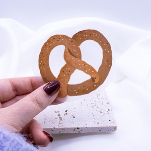 May include: A brown pretzel with a glossy finish and a sprinkle of salt on top. The pretzel is on a white background.