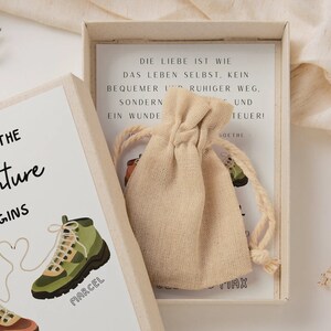 May include: A small, white box with a beige fabric bag inside. The box has a white card with the text "AND SO THE Adventure BEGINS" and two cartoon hiking boots with the names "MARCEL" and "TIAX" written on them. The card inside the box has German text that reads "DIE LIEBE IST WIE DAS LEBEN SELBST, KEIN BEQUEMER RUHIGER WEG, SONDERN EIN WUNDER UND TEUER!"