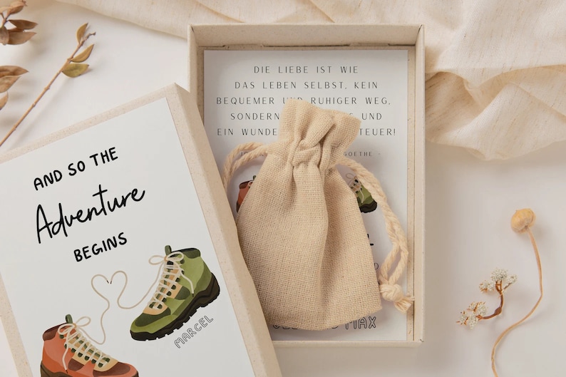 May include: A small, white box with a beige fabric bag inside. The box has a white card with the text "AND SO THE Adventure BEGINS" and two cartoon hiking boots with the names "MARCEL" and "TIAX" written on them. The card inside the box has German text that reads "DIE LIEBE IST WIE DAS LEBEN SELBST, KEIN BEQUEMER RUHIGER WEG, SONDERN EIN WUNDER UND TEUER!"