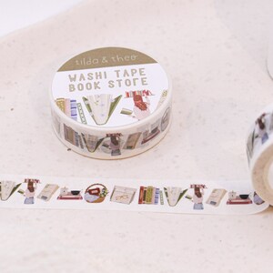 May include: A roll of washi tape with a book store theme. The tape features illustrations of books, flowers, and a person. The text on the tape reads "Washi Tape Book Store" and "Tilda & thee".