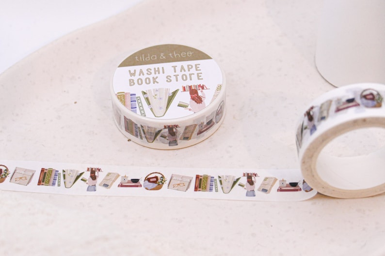 May include: A roll of washi tape with a book store theme. The tape features illustrations of books, flowers, and a person. The text on the tape reads "Washi Tape Book Store" and "Tilda & thee".
