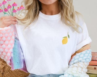 May include: White t-shirt featuring a small, yellow lemon illustration with a green leaf. The shirt is made of a soft, white fabric. The lemon design is located on the left chest area of the shirt.