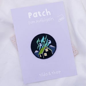 Peut inclure: Un patch thermocollant noir et vert avec une maison et une aurore boréale verte. Le texte "Patch zum Aufbügeln" et "tilda & theo" sont sur la carte.