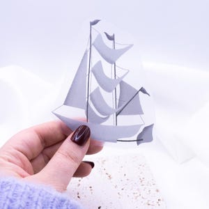 Könnte beinhalten: Ein weißer Papierschnitt eines Segelboots mit grauen Segeln. Das Segelboot wird in einer Hand mit dunkelrotem Nagellack gehalten.