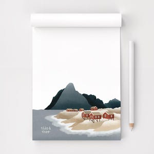 Könnte beinhalten: Ein Notizblock mit einer Küstenillustration. Das Kunstwerk zeigt rote Häuser an einem Sandstrand, mit einem dunklen Berg im Hintergrund. Der Text "tilda & theo" befindet sich unten links. Ein weißer Bleistift liegt rechts.