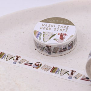 May include: A roll of washi tape with a book store theme. The tape features illustrations of books, glasses, and a person reading. The label on the roll reads "Washi Tape Book Store" and "tilda & theo".