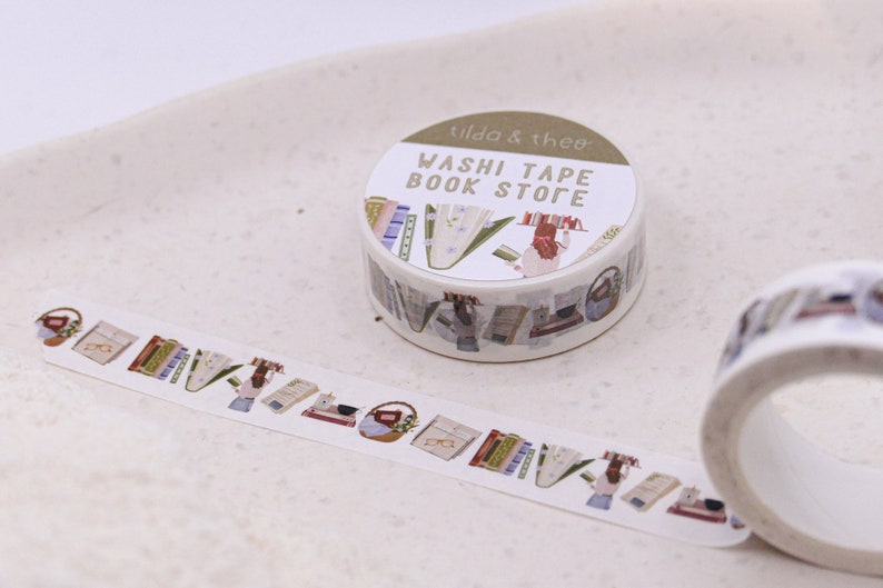 May include: A roll of washi tape with a book store theme. The tape features illustrations of books, glasses, and a person reading. The label on the roll reads "Washi Tape Book Store" and "tilda & theo".