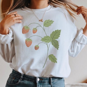 May include: White sweatshirt with a graphic of a strawberry plant with green leaves and red strawberries.