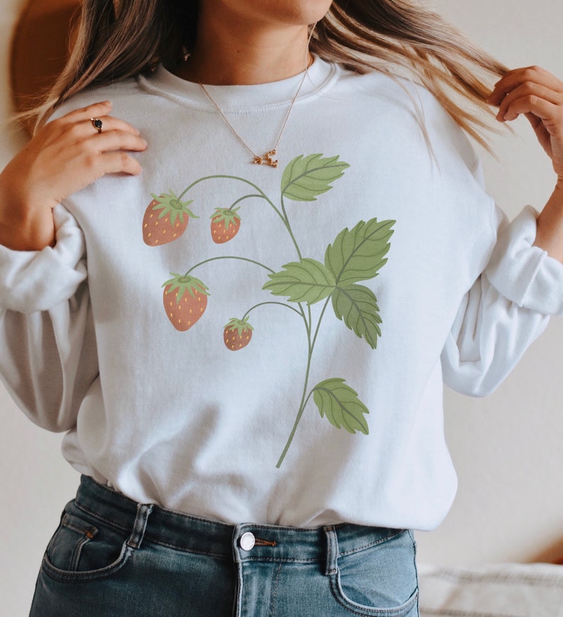 May include: White sweatshirt with a graphic of a strawberry plant with green leaves and red strawberries.