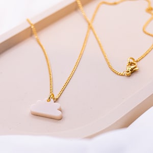 Peut inclure: Un collier en chaîne dorée avec un pendentif en forme de nuage blanc. Le collier est sur une surface brun clair.