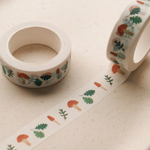 May include: Two rolls of white washi tape with a repeating pattern of red mushrooms and green leaves. One roll is unwinding, showing the pattern.