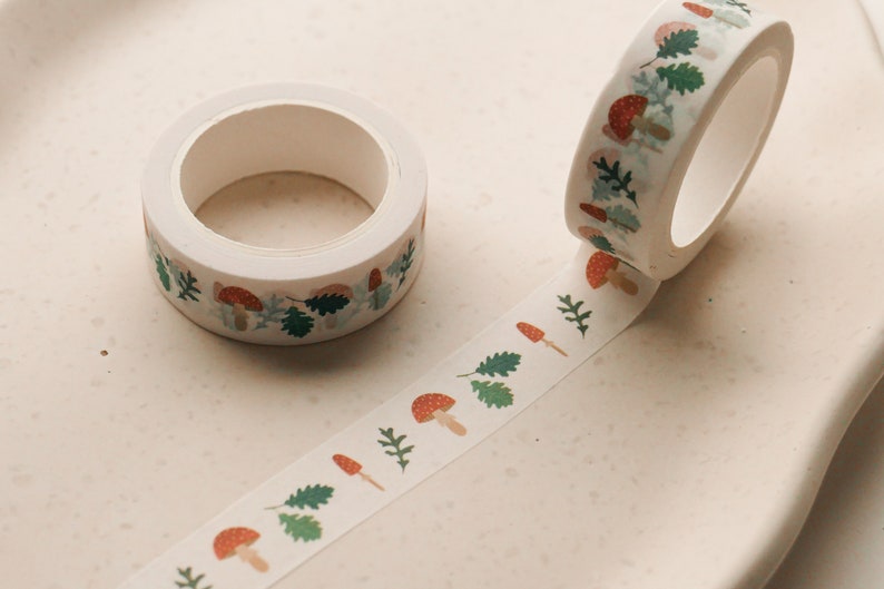 May include: Two rolls of white washi tape with a repeating pattern of red mushrooms and green leaves. One roll is unwinding, showing the pattern.