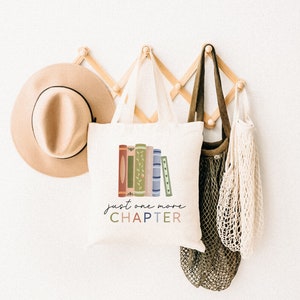 May include: A white tote bag with a stack of colorful books and the text "just one more CHAPTER" in a whimsical font. The bag is hanging on a wooden rack with a brown hat and a green and white string bag.