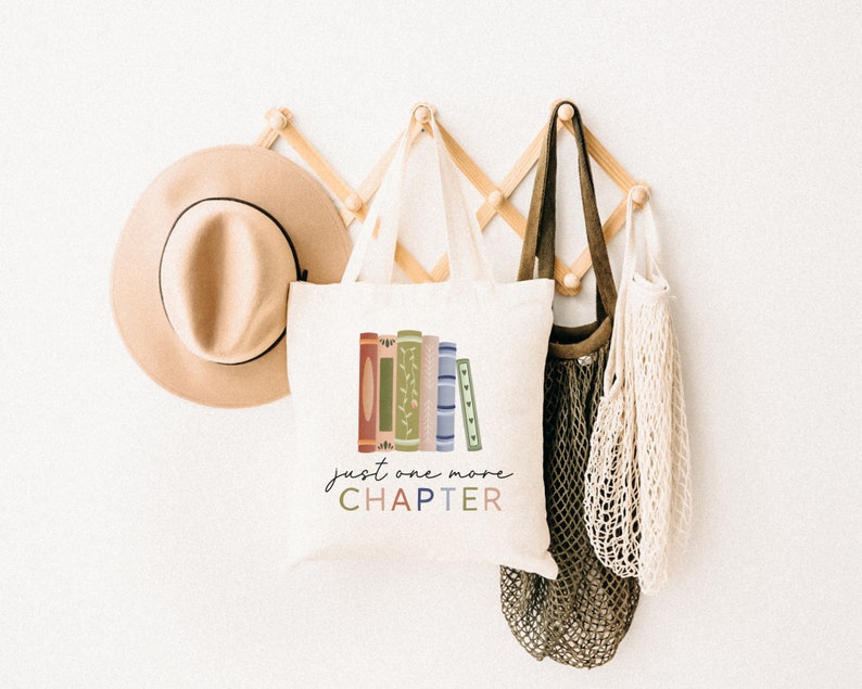May include: A white tote bag with a stack of colorful books and the text "just one more CHAPTER" in a whimsical font. The bag is hanging on a wooden rack with a brown hat and a green and white string bag.