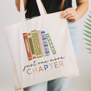 May include: A white canvas tote bag with a colorful illustration of stacked books and the text "just one more CHAPTER".