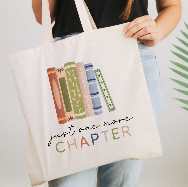 May include: A white canvas tote bag with a colorful illustration of stacked books and the text "just one more CHAPTER".