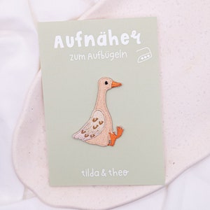 Könnte beinhalten: Bügeleisen-Aufnäher mit einer niedlichen Cartoon-Gans mit rosa Körper und orangefarbenen Füßen. Der Aufnäher befindet sich auf einer grünen Karte mit dem Text "Aufnäher zum Aufbügeln" und "tilda & theo".