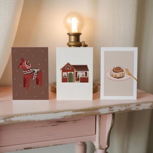 Puede incluir: Tres tarjetas de felicitación ilustradas sobre una mesa rosa. Las tarjetas presentan un caballo Dala rojo, una casa roja con una puerta verde y un rollo de canela con un tenedor dorado. Una lámpara vintage con una bombilla encendida está en el fondo.