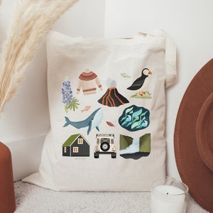 May include: A white canvas tote bag with a colorful illustration of Icelandic landmarks, including a whale, a puffin, a volcano, a house, a car, and lupine flowers. The text "Iceland" is printed on the bag.