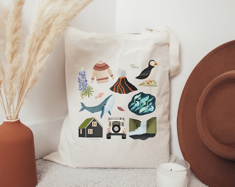 May include: A white canvas tote bag with a colorful illustration of Icelandic landmarks, including a whale, a puffin, a volcano, a house, a car, and lupine flowers. The text "Iceland" is printed on the bag.