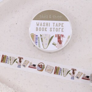 May include: A roll of washi tape with a book store theme. The tape features illustrations of books, flowers, and a person reading. The tape dispenser is labelled "Washi Tape Book Store" and "Tilda & Theo".
