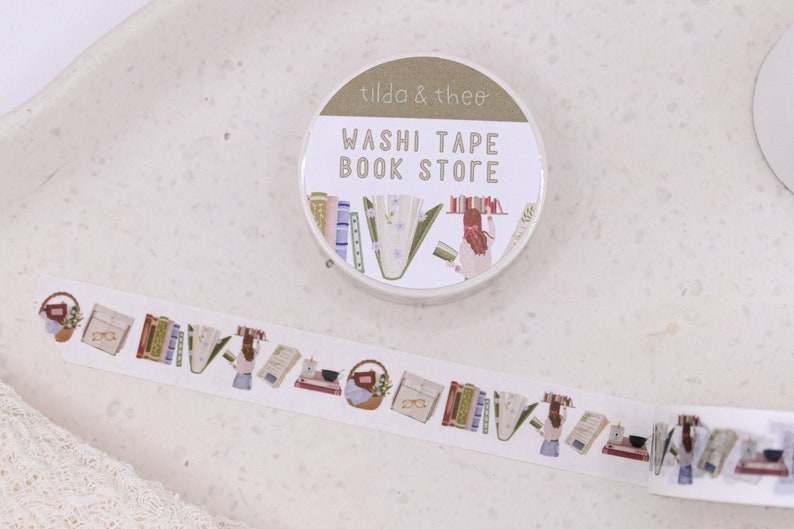 May include: A roll of washi tape with a book store theme. The tape features illustrations of books, flowers, and a person reading. The tape dispenser is labelled "Washi Tape Book Store" and "Tilda & Theo".