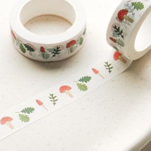 May include: White washi tape with a repeating pattern of red and white mushrooms and green leaves. The tape is rolled up on a white background.