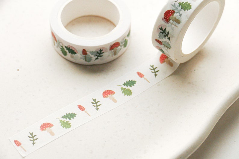 May include: White washi tape with a repeating pattern of red and white mushrooms and green leaves. The tape is rolled up on a white background.