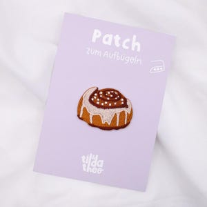 Könnte beinhalten: Gestickter Aufnäher eines Zimtschnecken-Brötchens mit weißer Glasur und braunen Streuseln, auf einer lila Karte. Die Karte trägt die Aufschrift "Patch" und "Zum Aufbügeln" mit einem Bügeleisensymbol. Die Marke "tilda theo" befindet sich unten.