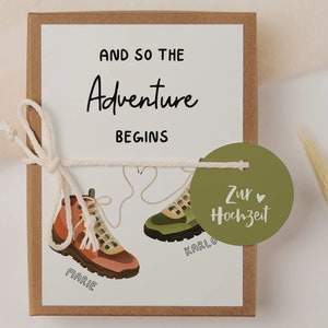May include: A white card with the text "And so the Adventure Begins" and two hiking boots with the names "Marie" and "Karlo" written on them. A green circle tag with the text "Zur Hochzeit" is attached to the card.