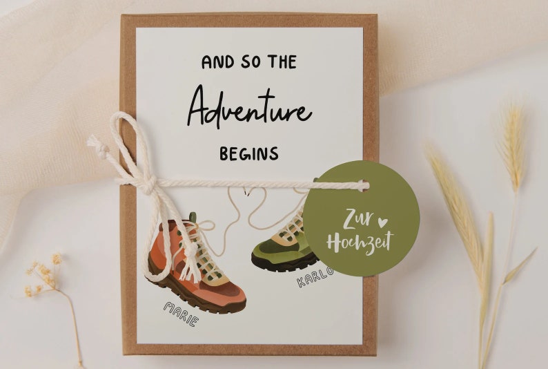 May include: A white card with the text "And so the Adventure Begins" and two hiking boots with the names "Marie" and "Karlo" written on them. A green circle tag with the text "Zur Hochzeit" is attached to the card.