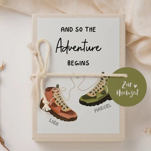 May include: A white gift box with a green and brown illustration of two hiking boots tied together with a white string. The box is wrapped with a white string and a green tag that says "Zur Hochzeit" with a heart. The text on the box says "And so the Adventure Begins" with the names "Lisa" and "Marcel" written on the boots.