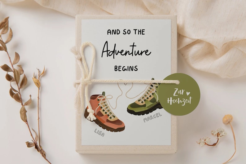 May include: A white gift box with a green and brown illustration of two hiking boots tied together with a white string. The box is wrapped with a white string and a green tag that says "Zur Hochzeit" with a heart. The text on the box says "And so the Adventure Begins" with the names "Lisa" and "Marcel" written on the boots.