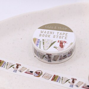 May include: A roll of washi tape with a book store theme. The tape features illustrations of books, a woman reading, and a basket of books. The tape is labelled "Washi Tape Book Store" and "Tilda & Theo".