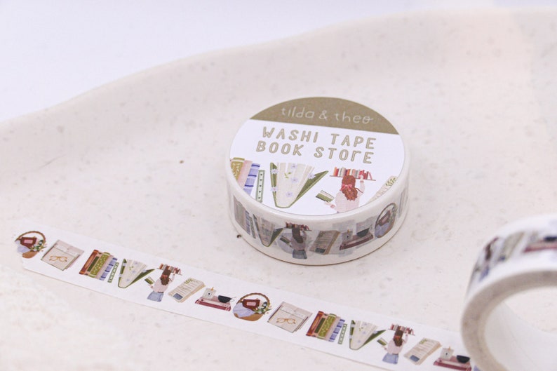 May include: A roll of washi tape with a book store theme. The tape features illustrations of books, a woman reading, and a basket of books. The tape is labelled "Washi Tape Book Store" and "Tilda & Theo".
