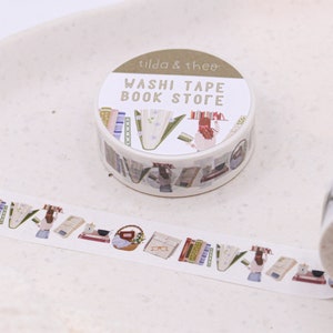 May include: A roll of washi tape with a book store theme. The tape features illustrations of books, a woman reading, and a basket of flowers. The label on the roll reads "Washi Tape Book Store" and "Tilda & Theo".