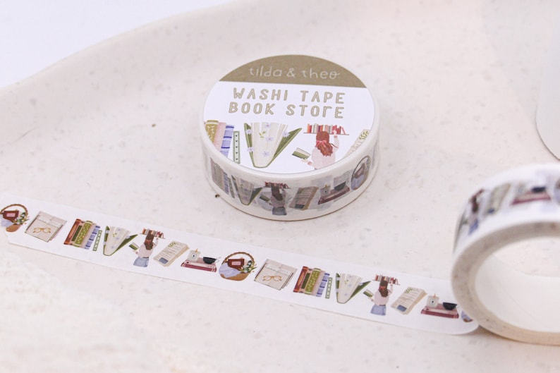 May include: A roll of washi tape with a book store theme. The tape features illustrations of books, a woman reading, and a basket of flowers. The label on the roll reads "Washi Tape Book Store" and "Tilda & Theo".
