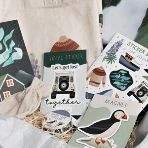 May include: A gift box filled with various items, including a tote bag, a vinyl sticker, a sticker sheet, a magnet, and a roll of washi tape. The items feature a variety of designs, including a house, a car, a puffin, and a sweater. The text on the sticker sheet reads "Sticker Sheet 009 Iceland".