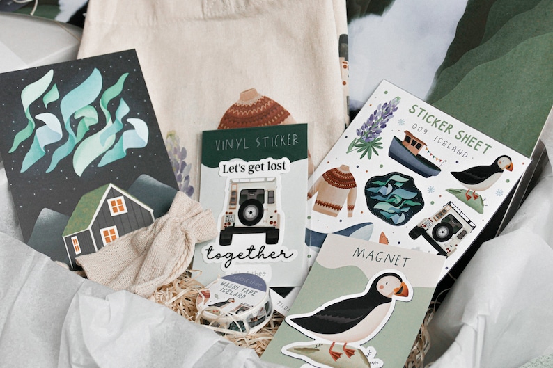 May include: A gift box filled with various items, including a tote bag, a vinyl sticker, a sticker sheet, a magnet, and a roll of washi tape. The items feature a variety of designs, including a house, a car, a puffin, and a sweater. The text on the sticker sheet reads "Sticker Sheet 009 Iceland".