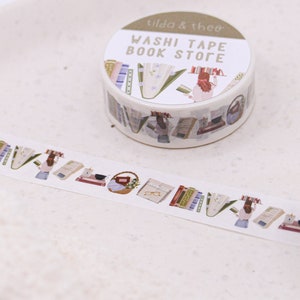 May include: A roll of white washi tape with a colourful design of books, glasses, and a person reading. The tape is labelled "Washi Tape Book Store" and "Tilda & Theo".