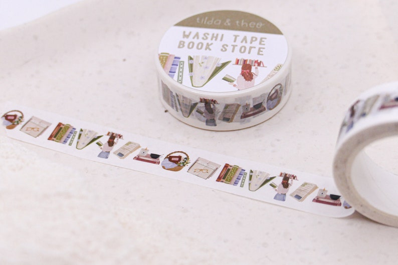 May include: A roll of white washi tape with a colourful design of books, glasses, and a person reading. The tape is labelled "Washi Tape Book Store" and "Tilda & Theo".