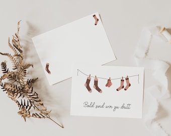 May include: Two white cards with a line of brown and pink socks hanging on a clothesline. The text on the card reads "Bald sind wir zu dritt".