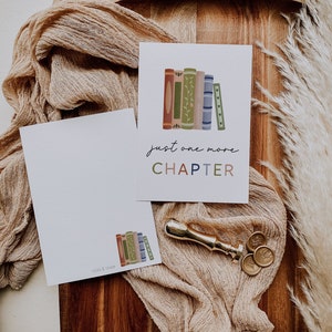 Peut inclure: Une carte blanche avec le texte "just one more CHAPTER" en lettres color&eacute;es. La carte pr&eacute;sente une illustration d'une pile de livres color&eacute;s avec des d&eacute;tails floraux. La carte est pos&eacute;e sur une surface en bois brun avec un morceau de tissu et un sceau de cire.