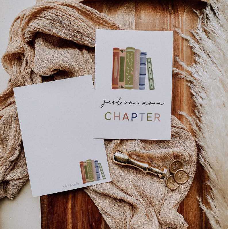 Peut inclure: Une carte blanche avec le texte "just one more CHAPTER" en lettres color&eacute;es. La carte pr&eacute;sente une illustration d'une pile de livres color&eacute;s avec des d&eacute;tails floraux. La carte est pos&eacute;e sur une surface en bois brun avec un morceau de tissu et un sceau de cire.
