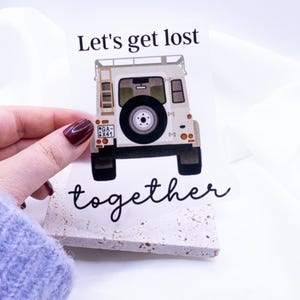Könnte beinhalten: Ein weißer Aufkleber mit einer Grafik eines beigefarbenen Geländewagens, einem Reserverad und einer Leiter. Der Text "Let's get lost together" ist in schwarzer Schrift geschrieben. Der Aufkleber wird von einer Hand mit weinrotem Nagellack gehalten.