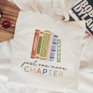 May include: A white canvas tote bag with a design of stacked books with floral details. The text "just one more CHAPTER" is printed in colorful letters below the books.